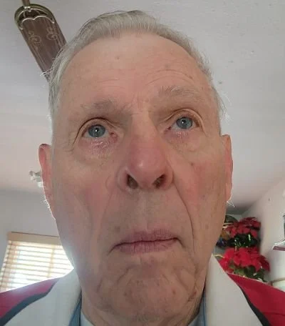 Close-up of an elderly man with gray hair, blue eyes, and a serious expression, indoors with a Christmas poinsettia plant and window in the background.