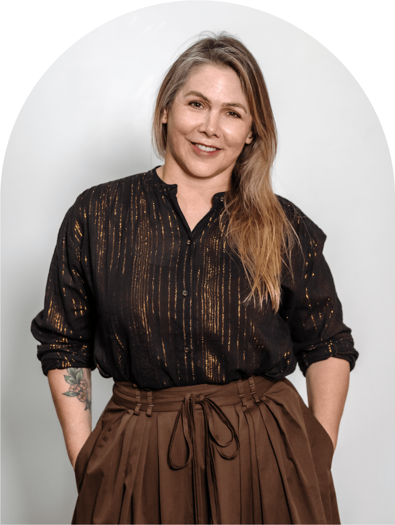 A woman with long, wavy blonde hair, wearing a black blouse with gold accents and brown pleated pants, standing against a plain white background.