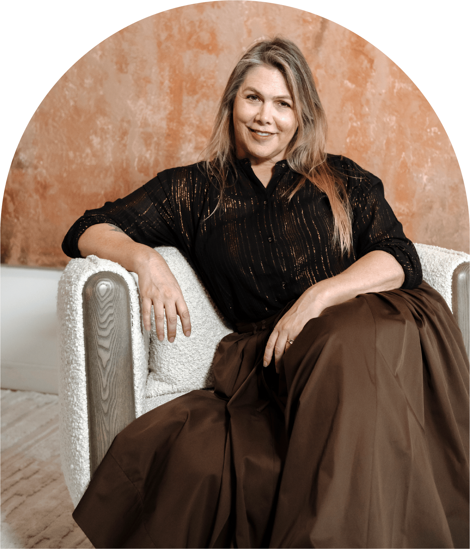 A woman with long blonde hair sitting on a white textured armchair against a terracotta-colored wall, wearing a black top and a brown skirt, smiling at the camera.