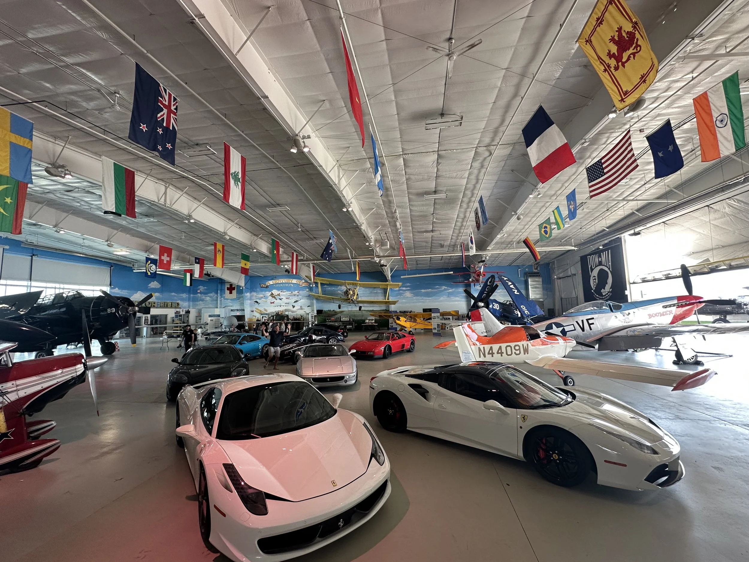 An indoor aviation museum with various aircraft and cars on display, flags hanging from the ceiling, and visitors admiring the exhibits.