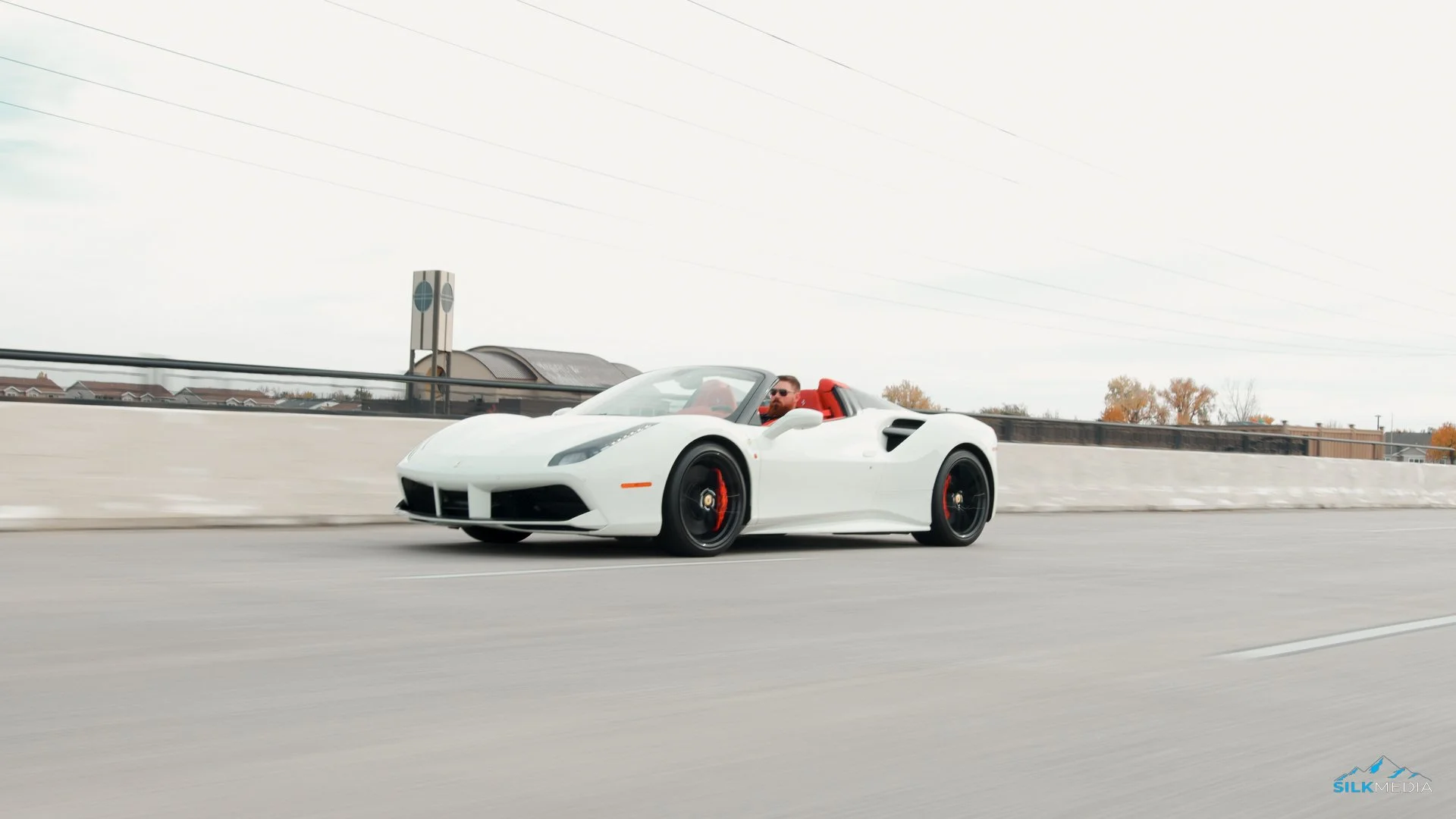 A white Lamborghini convertible sports car with a red interior driving on a highway, driven by a man with sunglasses and a beard. The background is overcast with some trees and buildings visible.
