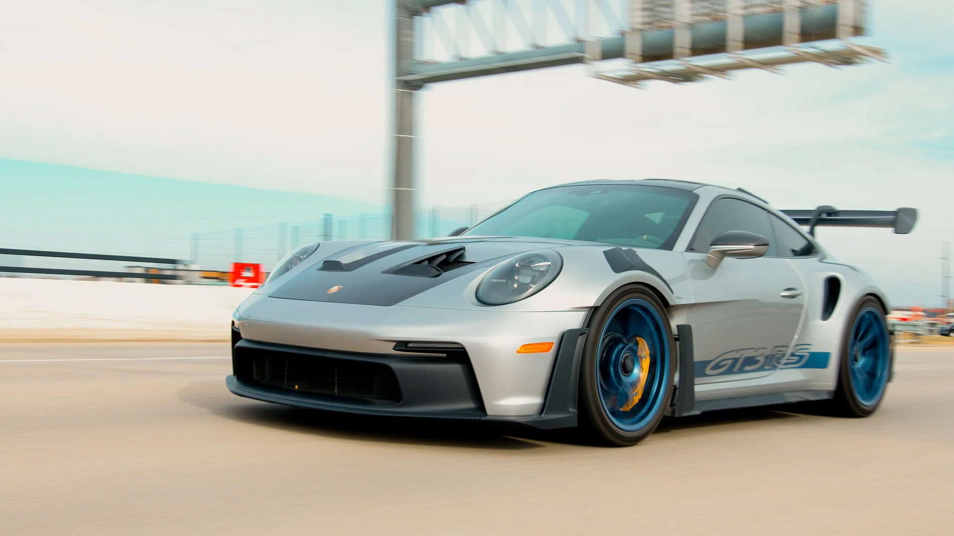 A silver Porsche 911 GT3 RS race car with blue wheels and a large rear wing, driving on a race track.