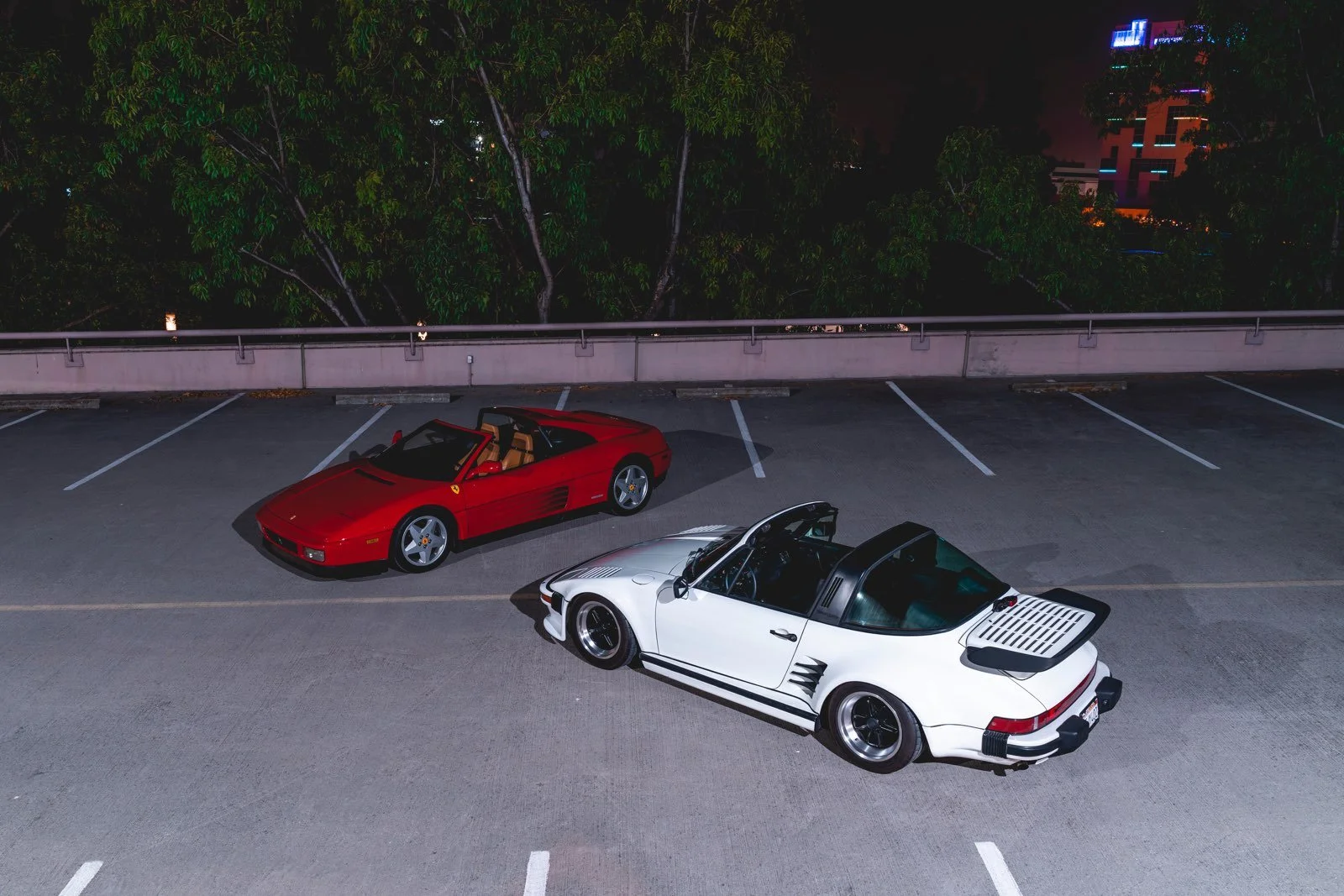 A red Ferrari and a white Porsche 911 with a black roof parked side by side in an empty parking lot at night.