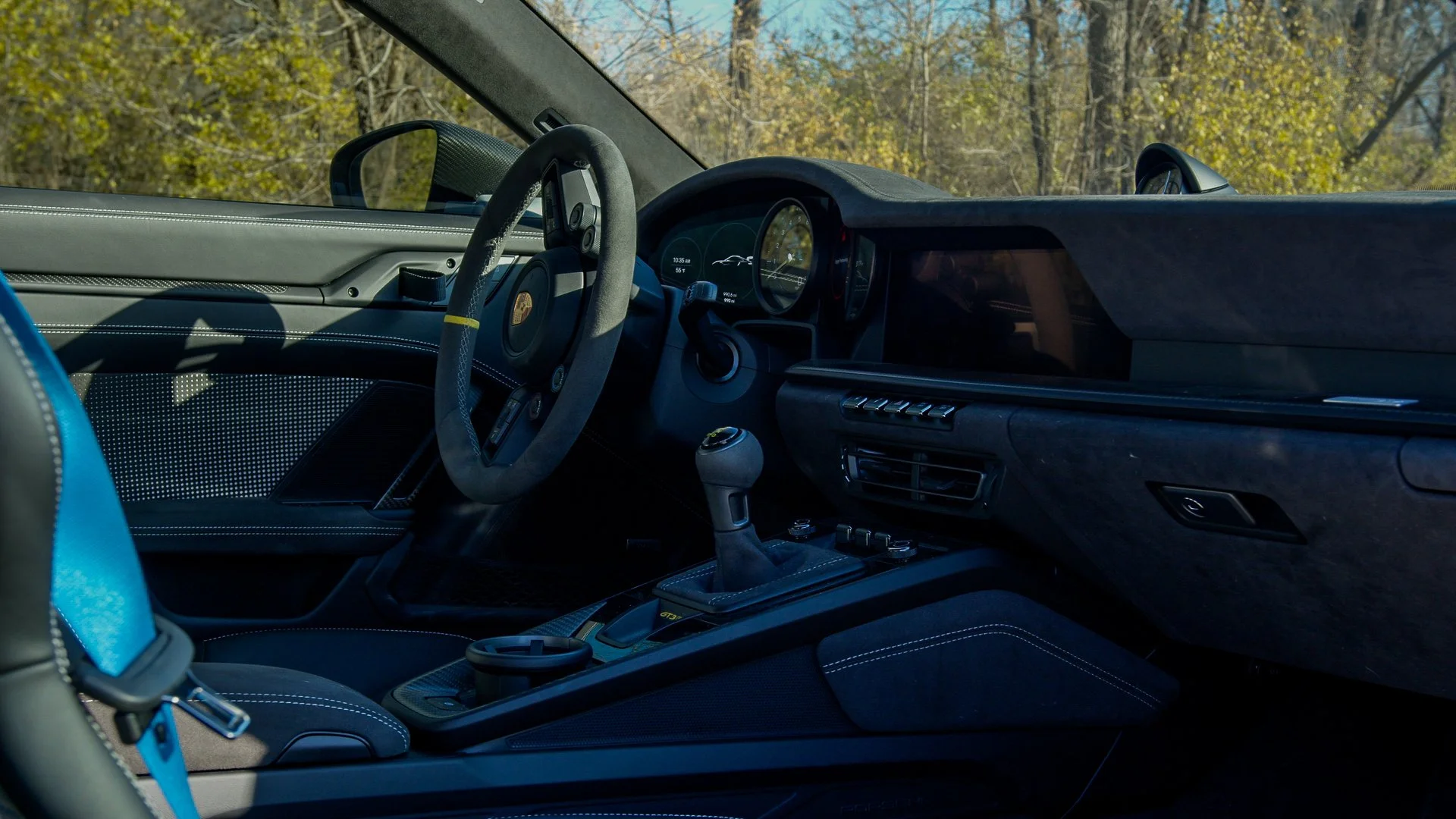 Interior view of a luxury sports car with a carbon fiber dashboard, racing-style steering wheel with a yellow stripe, manual gear shifter, digital instrument cluster, and a blue racing seatbelt. Outside, trees and a clear sky are visible.