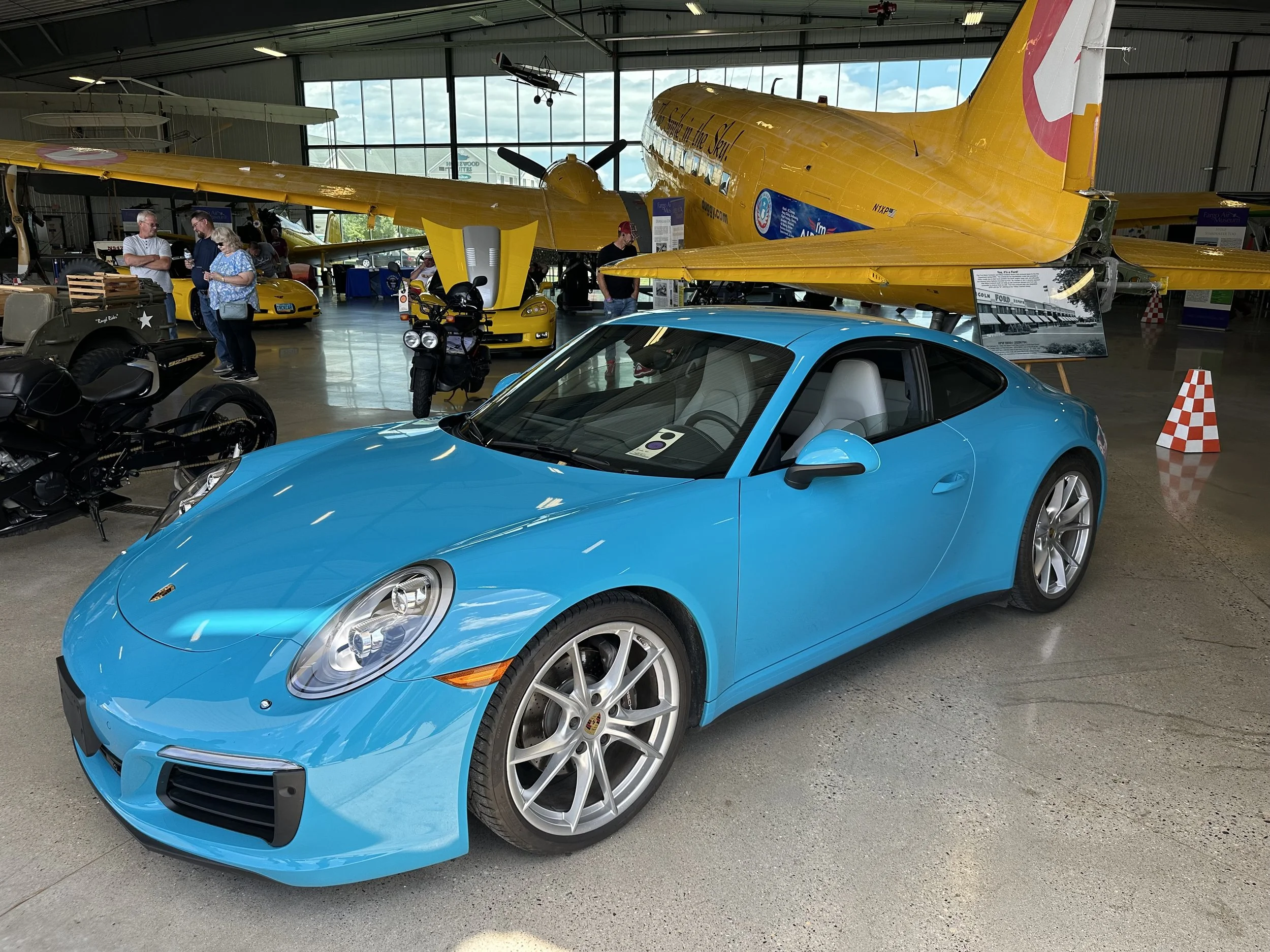 Blue Porsche sports car displayed in an aviation museum with vintage planes and people in the background.