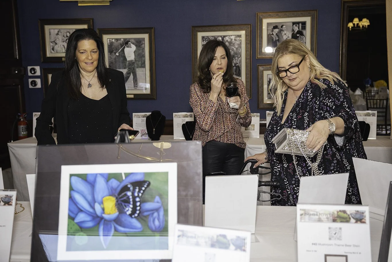Three women browsing jewelry and art displays at an indoor event, with framed photographs on the wall behind them.