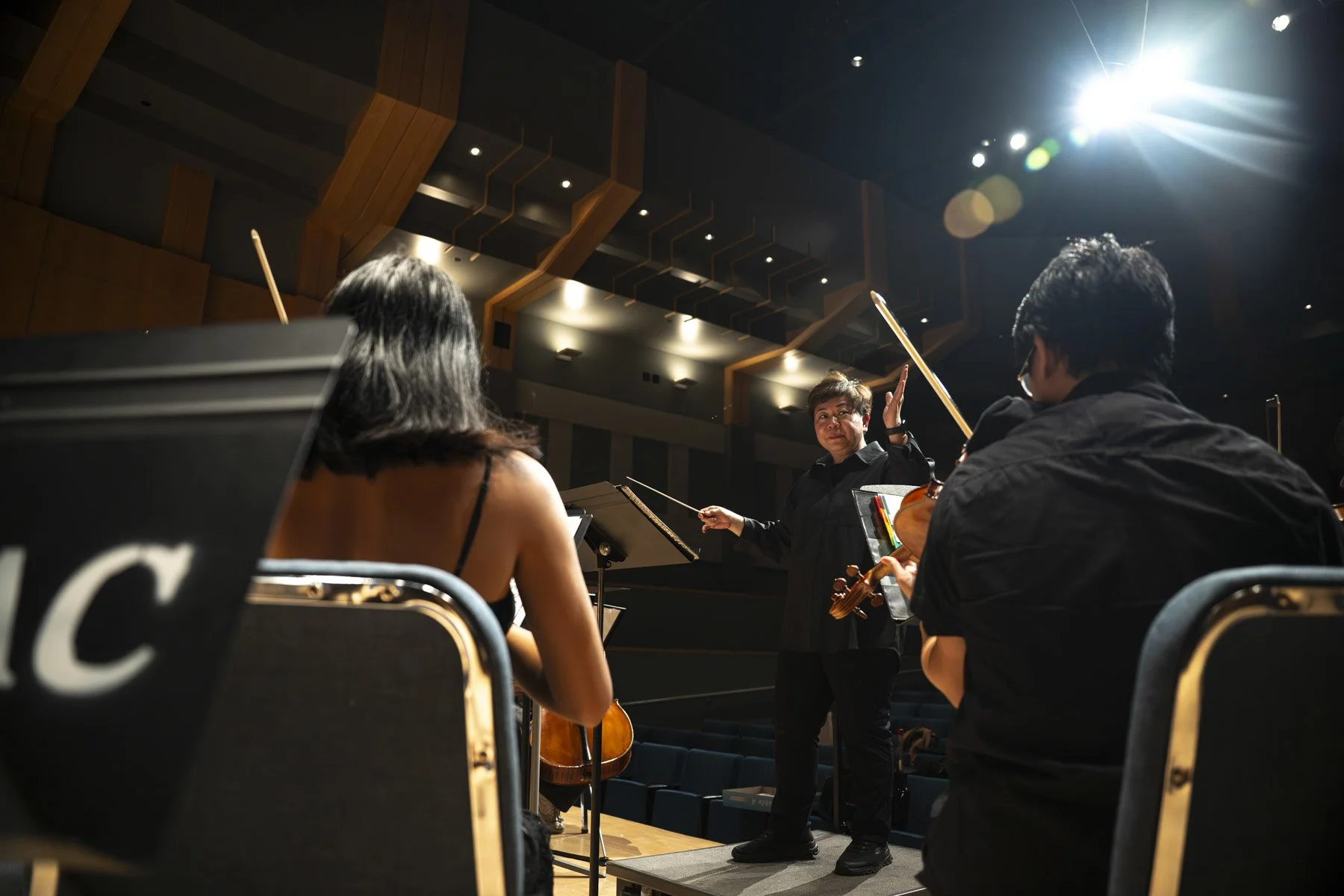 Conductor leading an orchestra during a rehearsal or performance in a concert hall, with musicians playing string instruments.