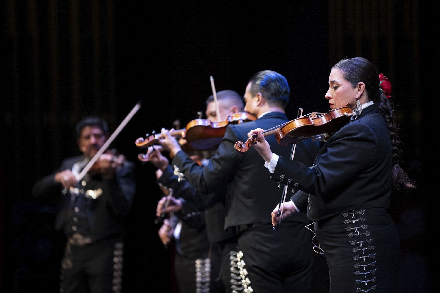 Mariachi musicians performing with violins and guitar on stage in traditional black outfits.