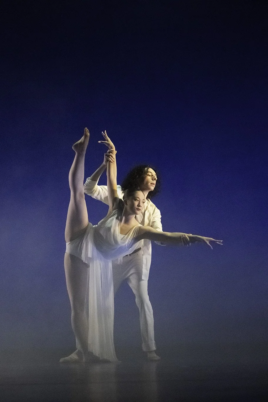 Two ballet dancers performing a duet on stage, with one dancer standing and the other in an arabesque position, against a dark blue background.