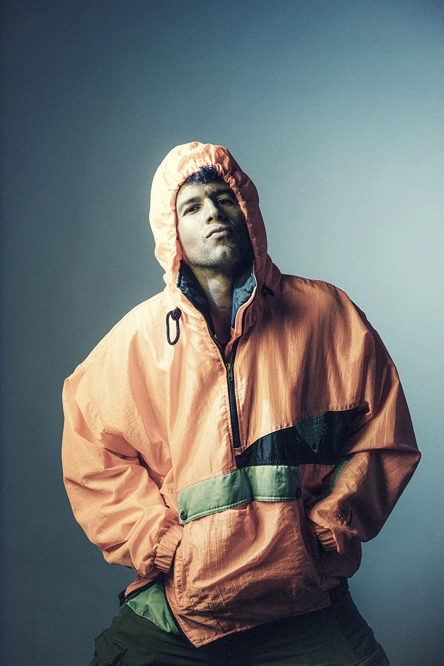 Man in a pink windbreaker with hood standing confidently against gradient teal background.