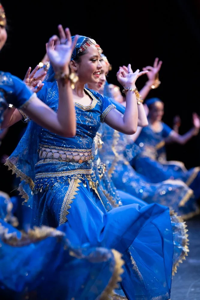 Group of women in traditional blue and gold dance costumes performing a dance on stage, smiling with hands raised.