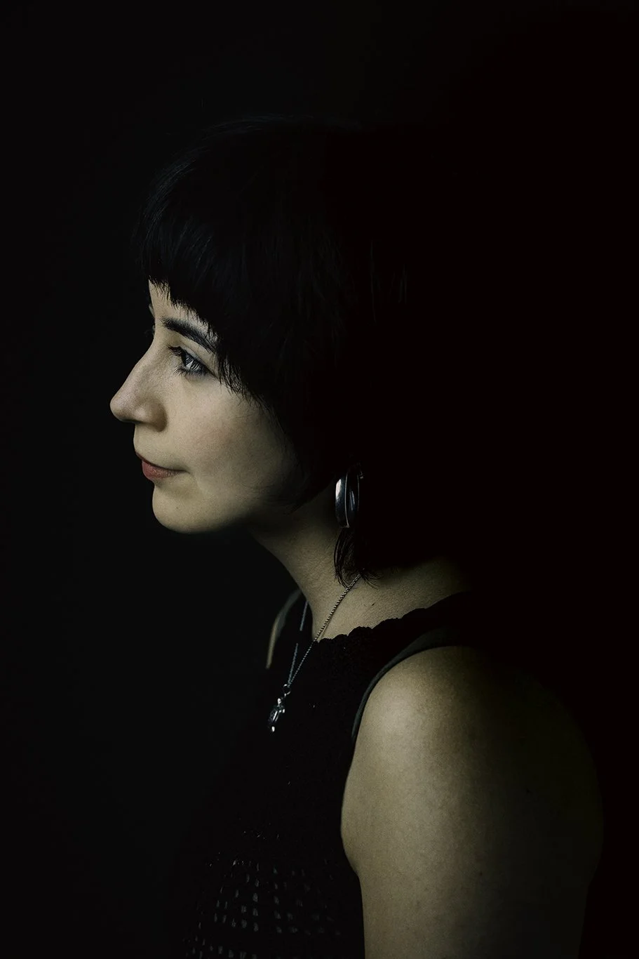 Profile of young woman with short black hair wearing black top, earrings, and necklace against dark background.