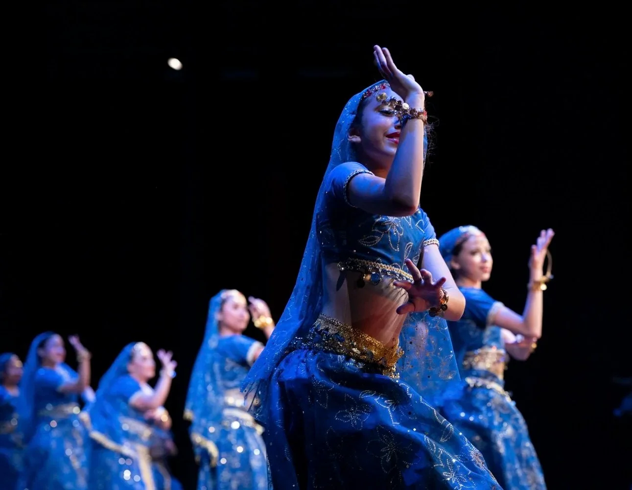 Young girls performing traditional dance on stage in blue and gold costumes under stage lighting.