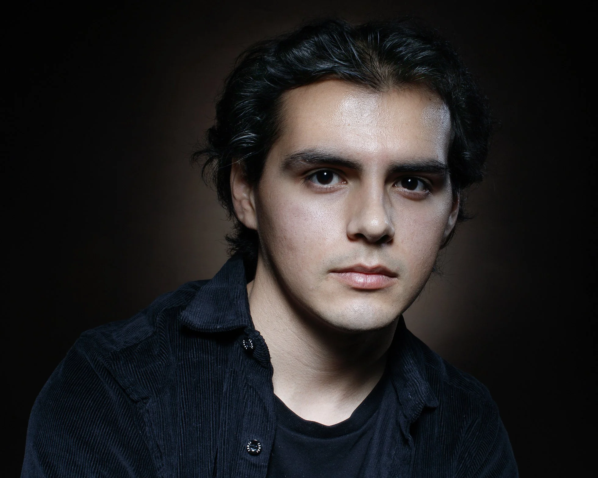 Portrait of young man with dark hair and fair skin wearing black shirt against dark background.