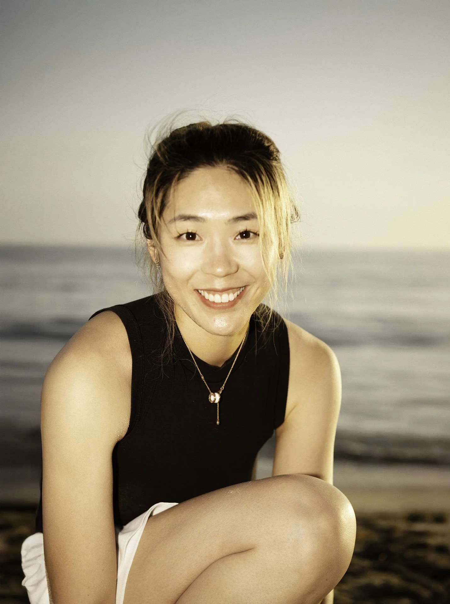 Young woman smiling while sitting on a beach during sunset with ocean in the background.
