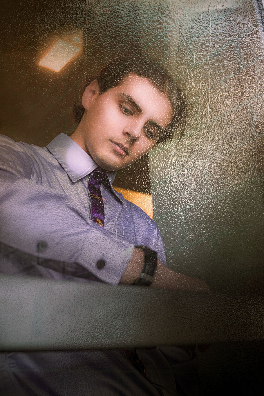 Young man with dark hair wearing button-up shirt and watch taking selfie in front of foggy window.