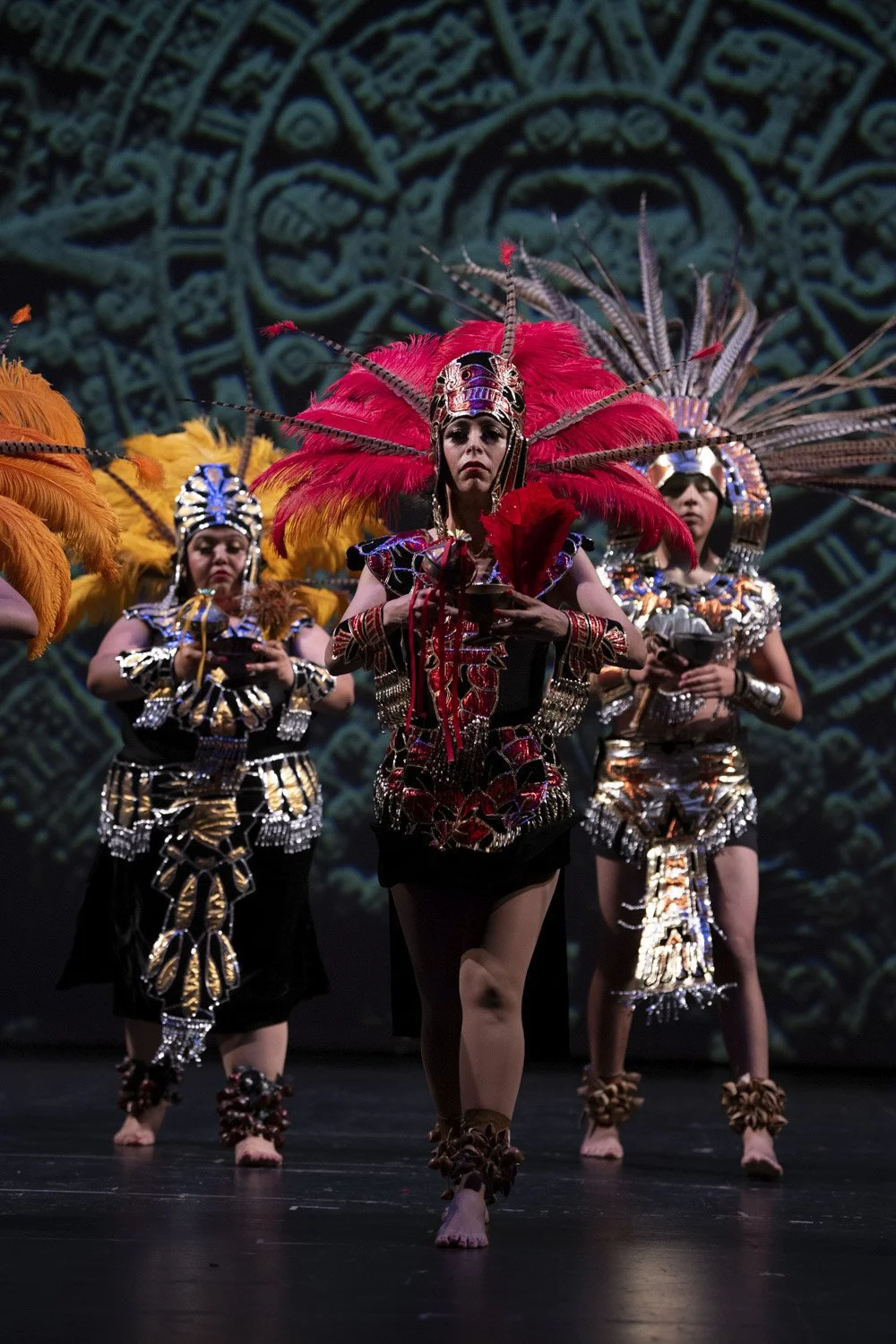 Group of performers in traditional, colorful costumes with feathered headdresses, dancing in front of a decorative background.