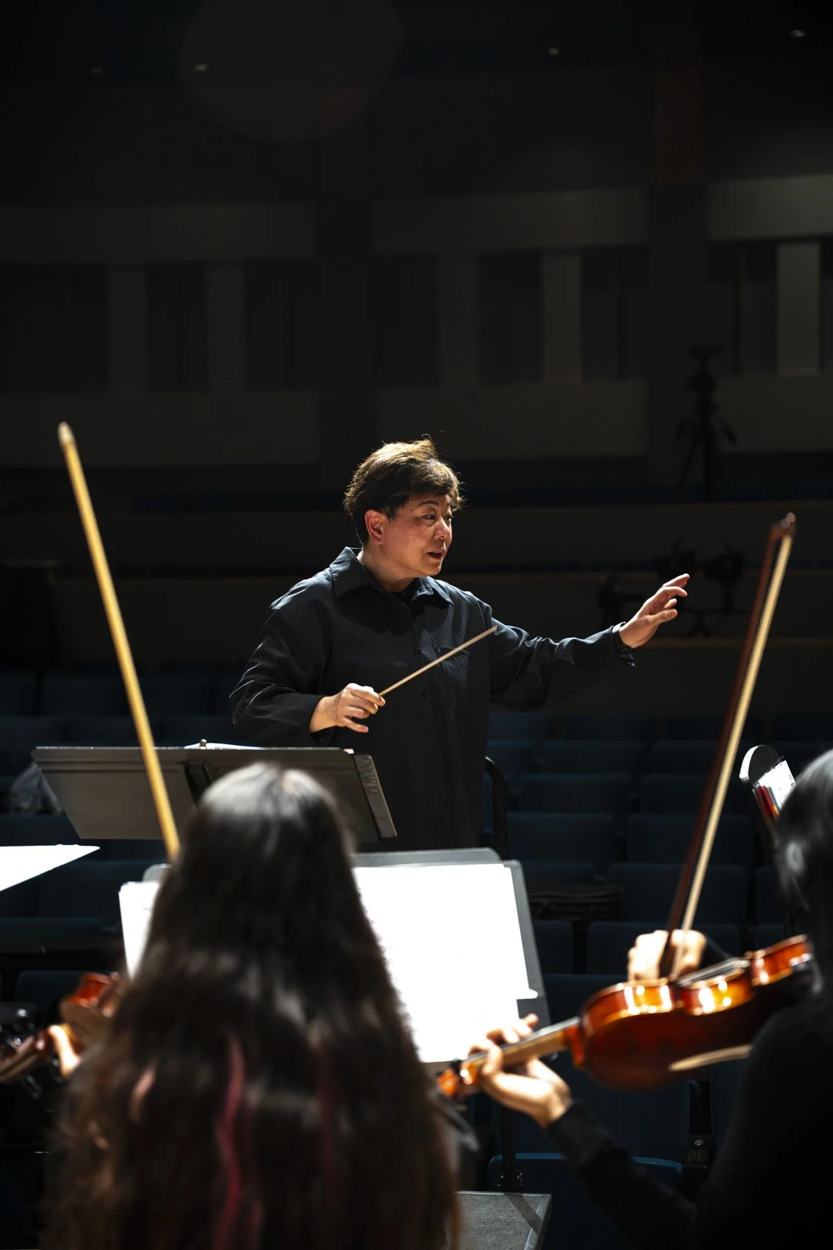 Woman conductor leading orchestra with violinists in dimly lit concert hall.