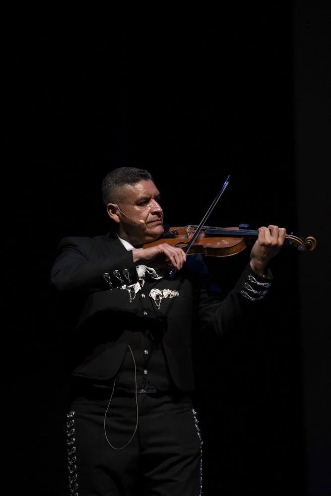 Man in traditional Mexican attire playing violin on stage with dark background.