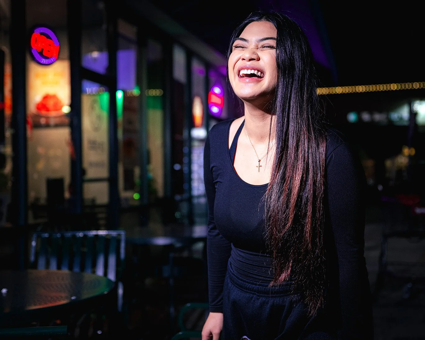 Woman with long dark hair laughing outdoors at night, illuminated by neon lights in front of restaurant or bar.