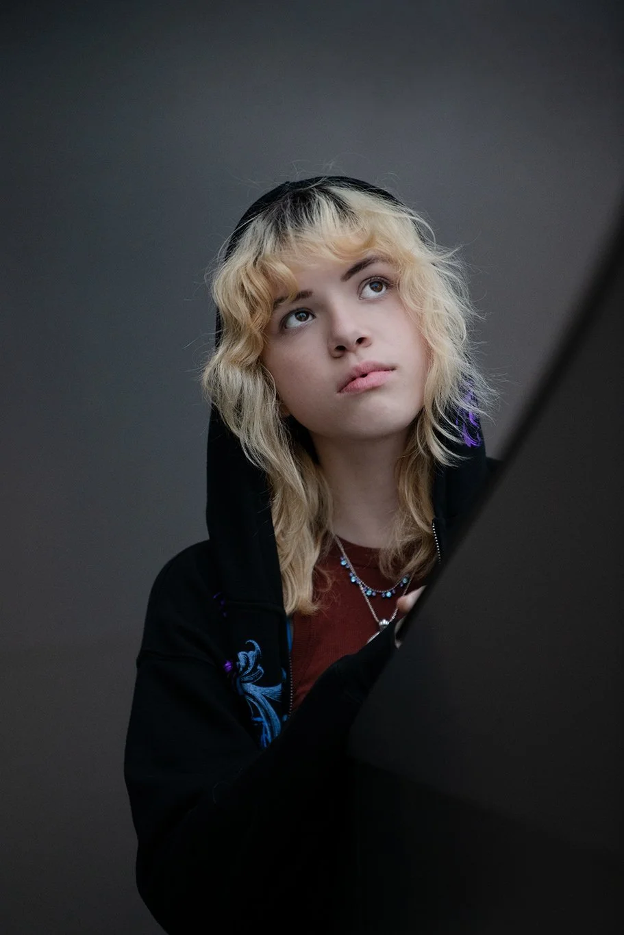 Young woman with blonde curly hair wearing a black hoodie with blue designs using a laptop thoughtfully.
