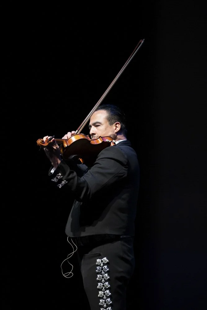 Man wearing a black tuxedo playing a violin on stage with a dark background.