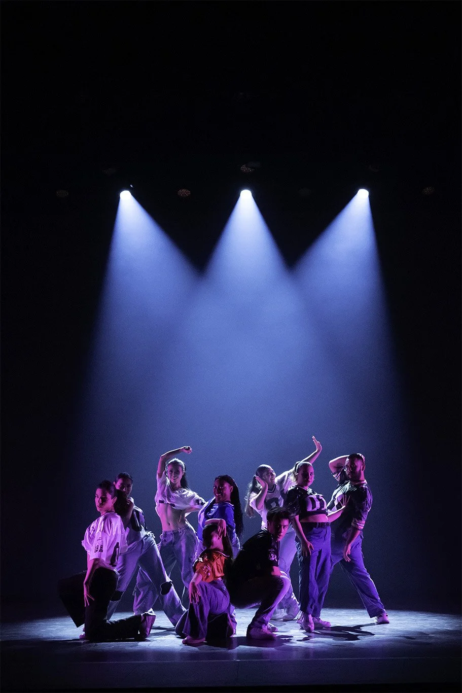 Group of dancers in casual streetwear performing on stage under blue spotlights.