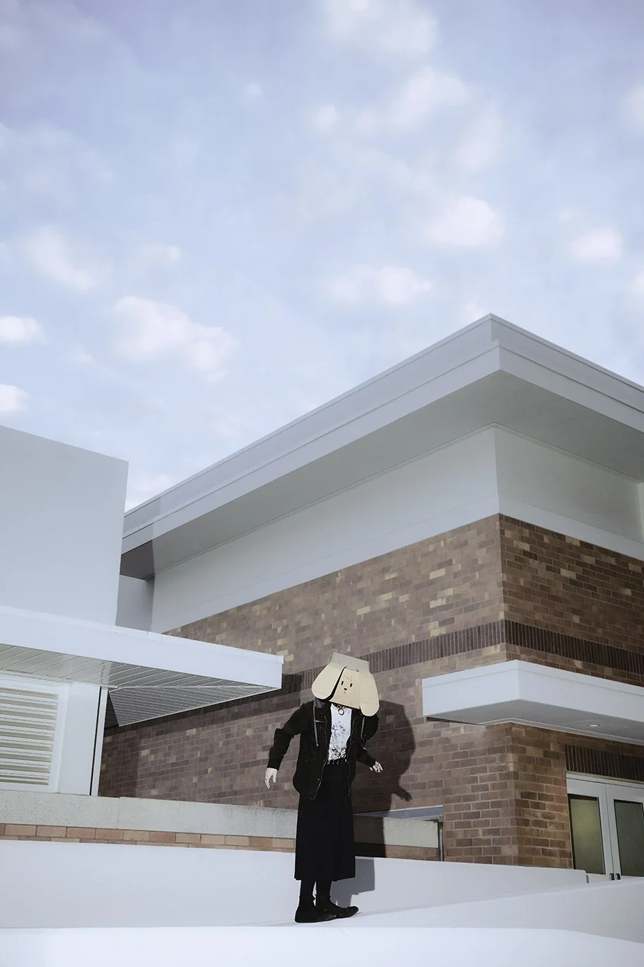 Person wearing cartoon dog head standing outside a modern red brick and white building under blue sky.