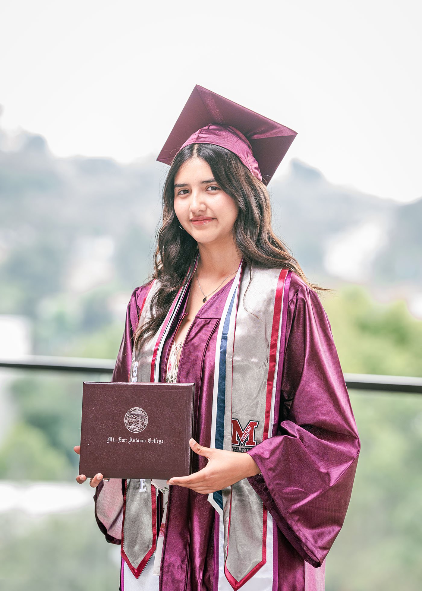 A young woman in a maroon graduation gown and cap holding a diploma from Mt. San Antonio College, standing outdoors with a blurred background of trees and cloudy sky.