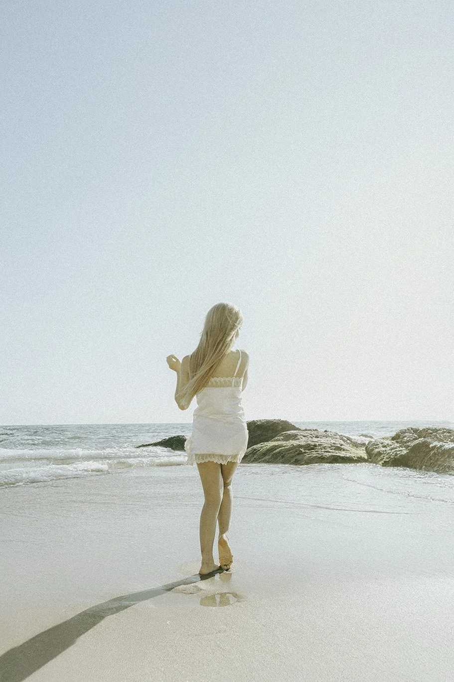 Woman with long blonde hair in a white dress walking on a beach with ocean and rocks in the background.