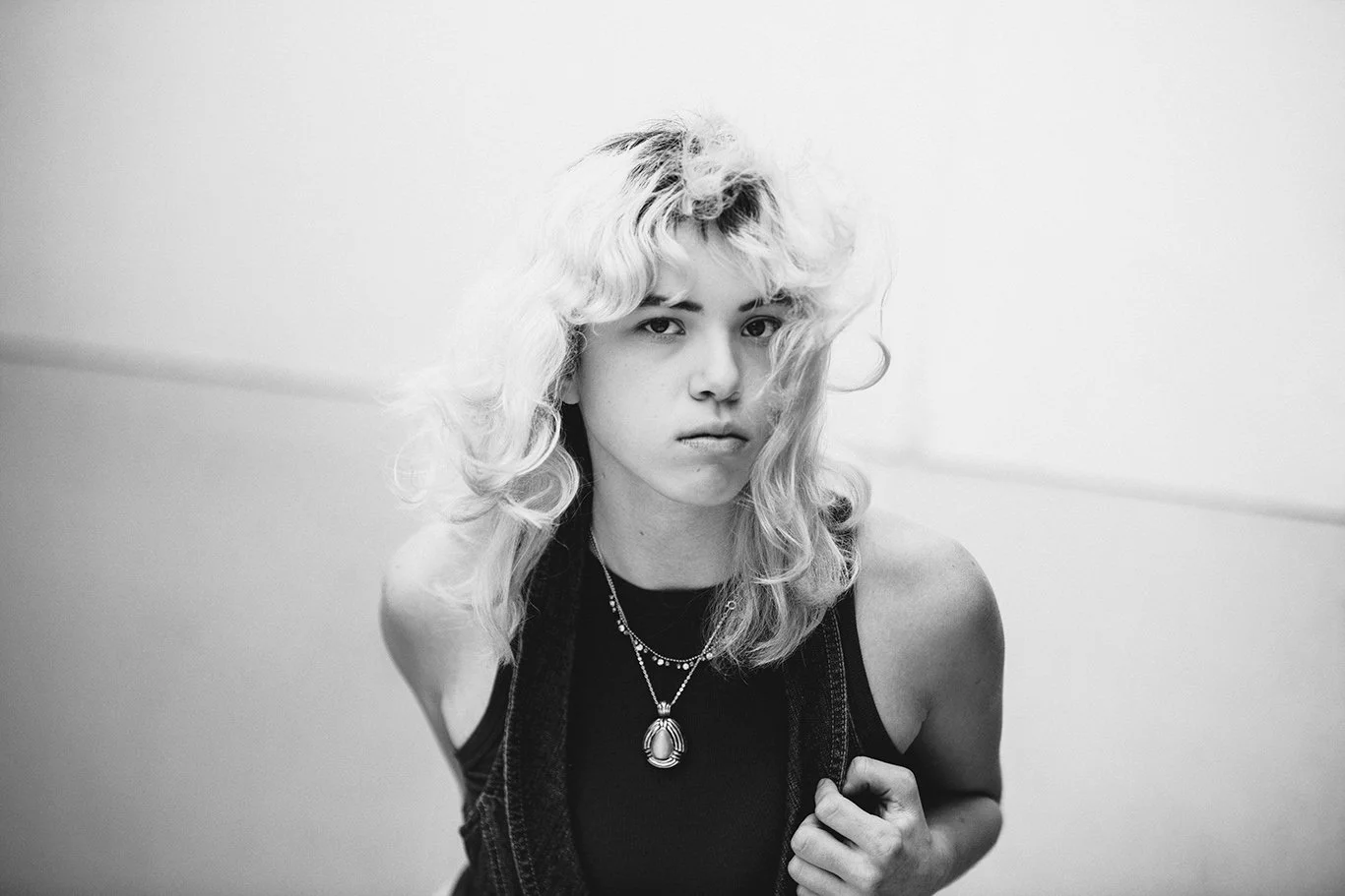 Black and white portrait of a young woman with wavy shoulder-length blonde hair wearing a sleeveless top and layered necklaces.