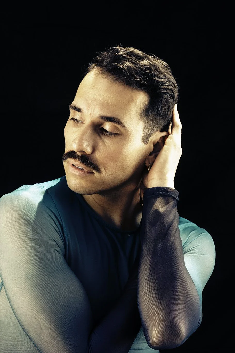 Portrait of a man with dark hair and mustache, wearing a blue long-sleeve shirt with black mesh sleeves, against a black background, looking down with eyes closed and hand behind his ear.
