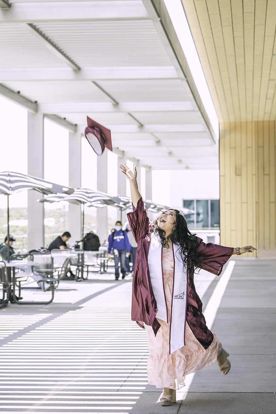 A woman in a maroon graduation gown and pink dress is celebrating outdoors, reaching up to toss her cap into the air with a joyful expression. There are other people in the background sitting and walking, and the setting appears to be at a modern bui