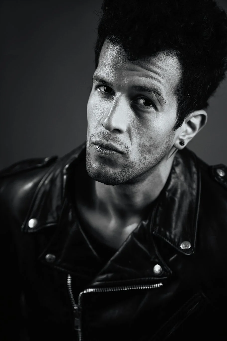 Black and white portrait of young man with curly hair in a leather jacket looking at the camera.