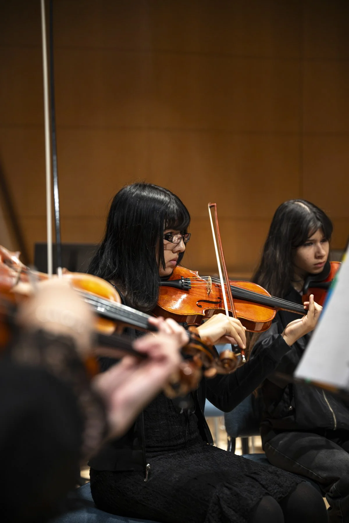 Two young women playing violins indoors with wooden walls.