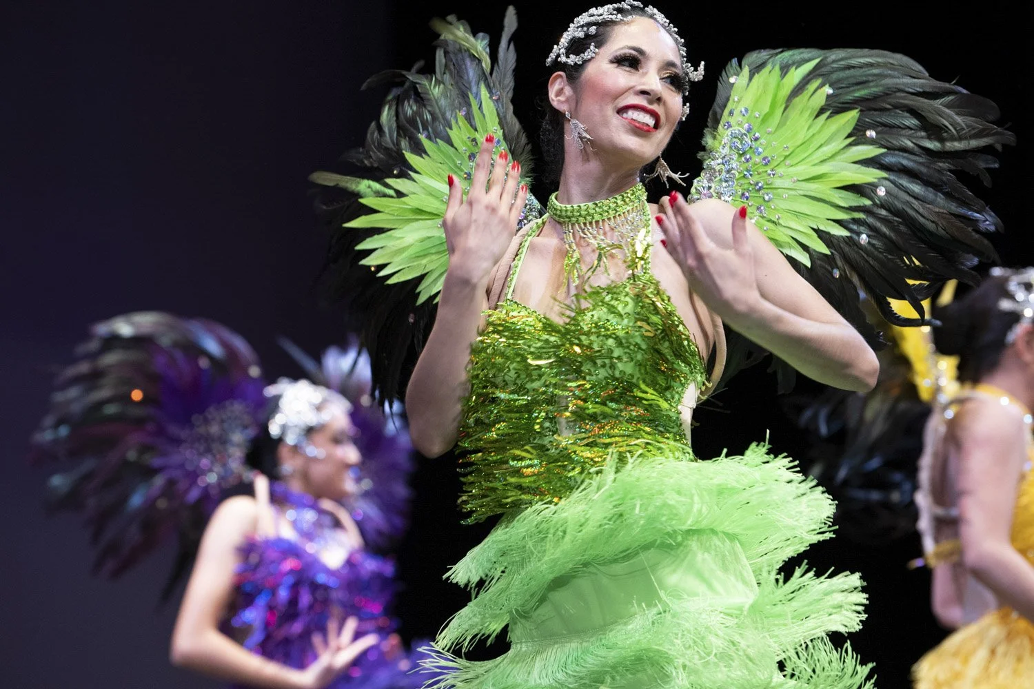 A woman wearing a bright green, feathered dress with matching jewelry, smiling and posing on stage during a costume performance, with other women in similar costumes in the background.