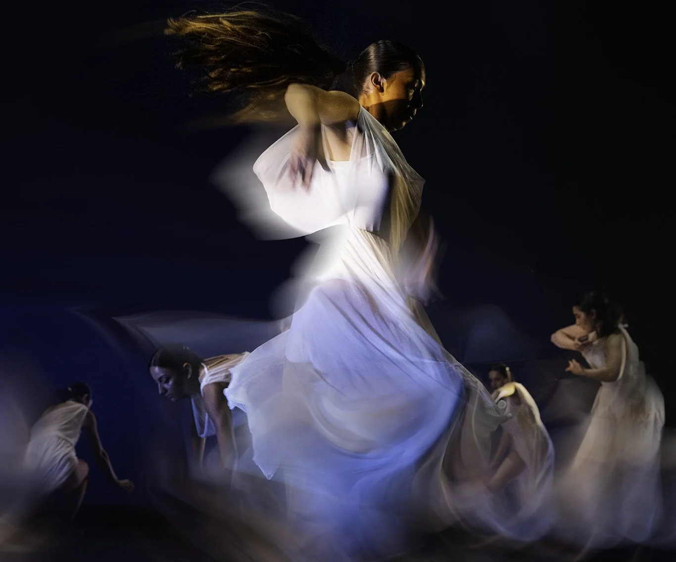 A woman in a white dress dancing with multiple blurred images around her, creating a motion effect against a dark background.