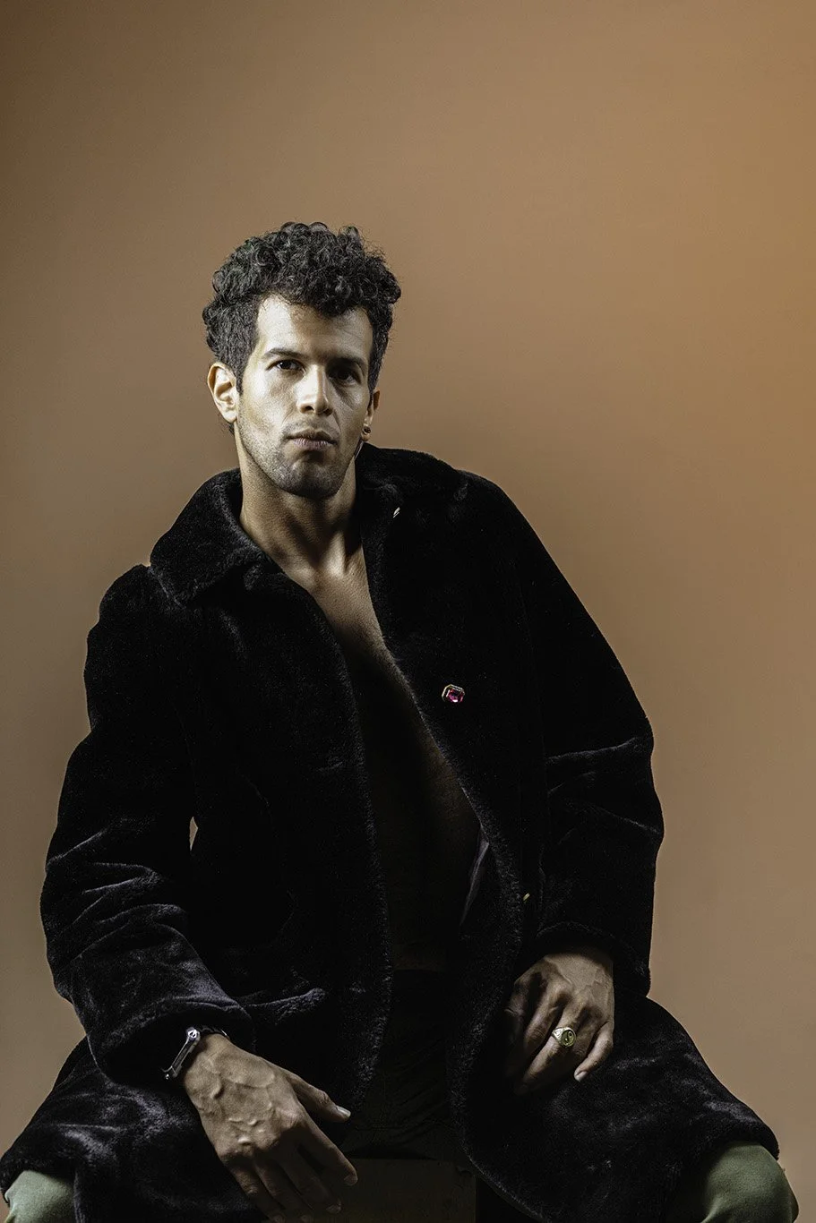 Young man with curly hair in black velvet coat holding a ring, sitting against brown background.