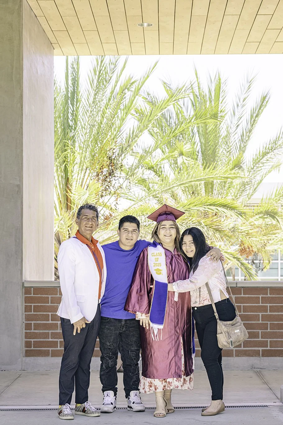 A young woman in a graduation gown and cap stands with three family members in front of palm trees. The woman wears a sash and stole that say "Class of 2025." The family members are standing closely, smiling and hugging, celebrating her graduation ou