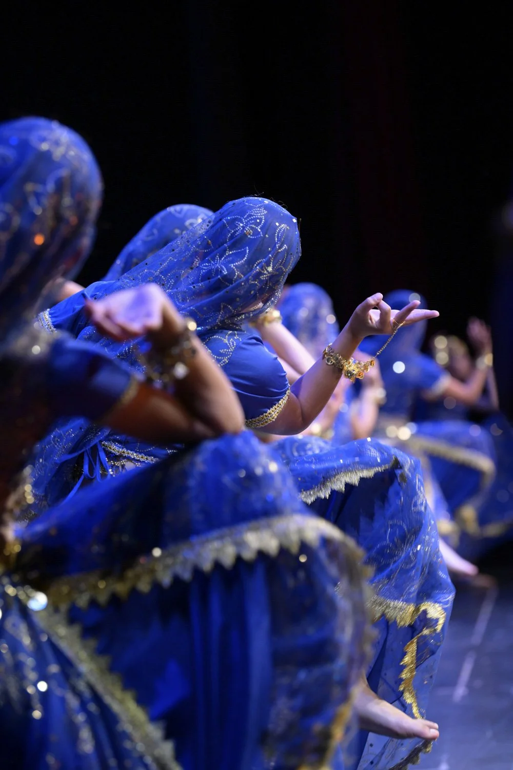 Women in blue traditional costumes with gold accents sitting and performing a dance or musical act.