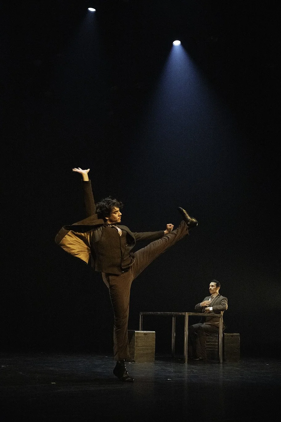 A person performing a dance or theatrical pose on stage with a man seated at a table in the background, under dramatic stage lighting.