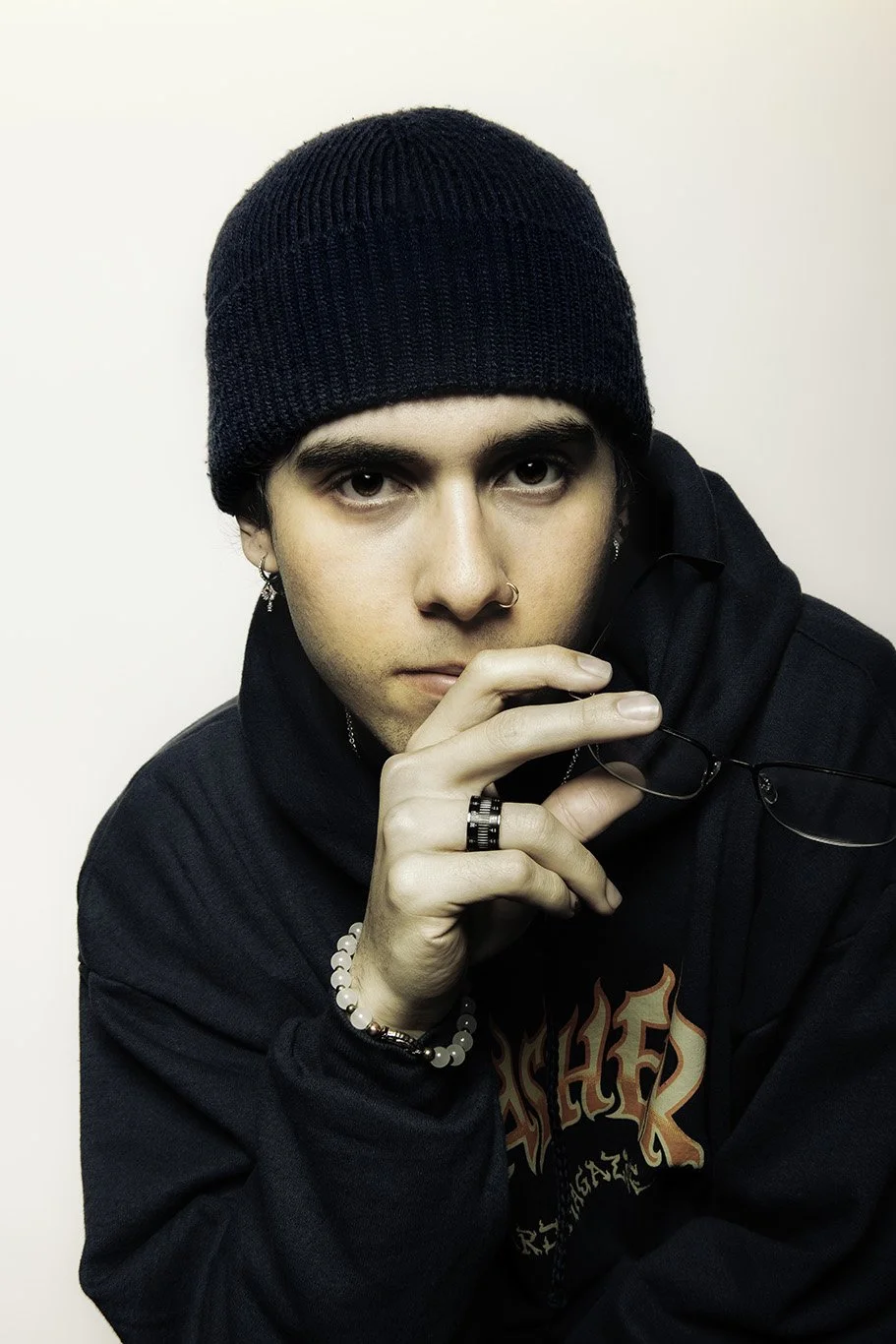 Young man with dark hair in black beanie and hoodie holding glasses near face looking at camera.