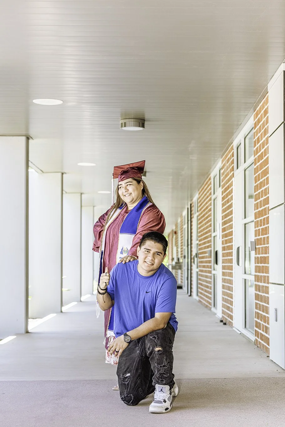 A graduate in a maroon cap and gown with a blue stole, smiling and standing behind a young man who is kneeling on one knee, wearing a blue T-shirt and ripped jeans.