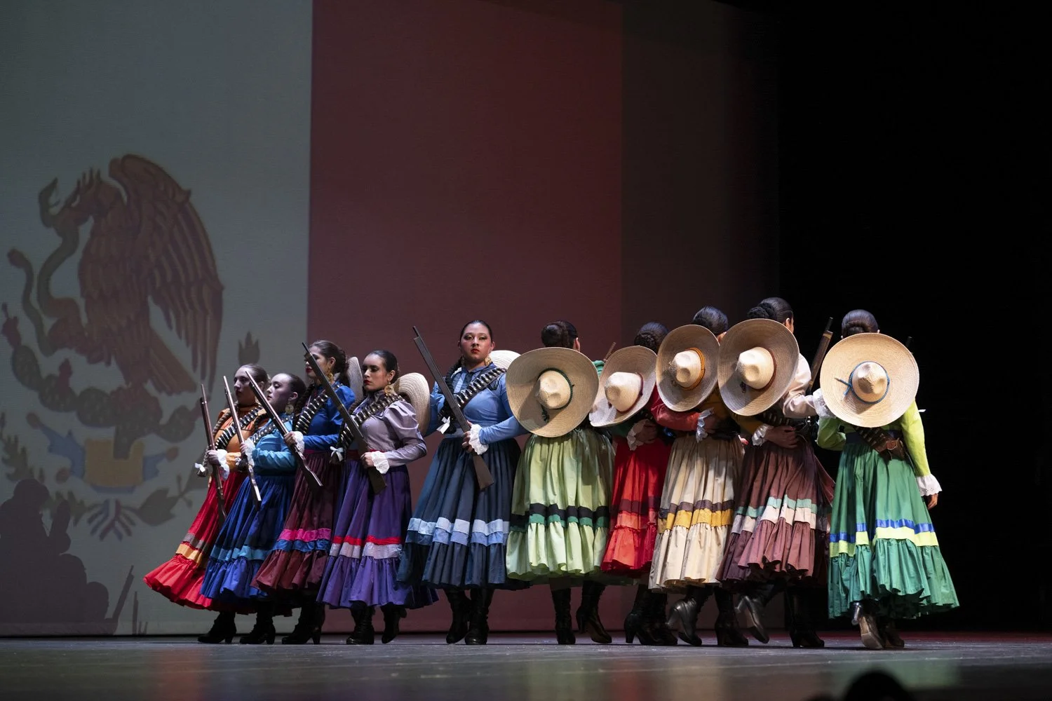 Mexican dance group performing in traditional dresses and large sombreros with Mexican flag projected behind.