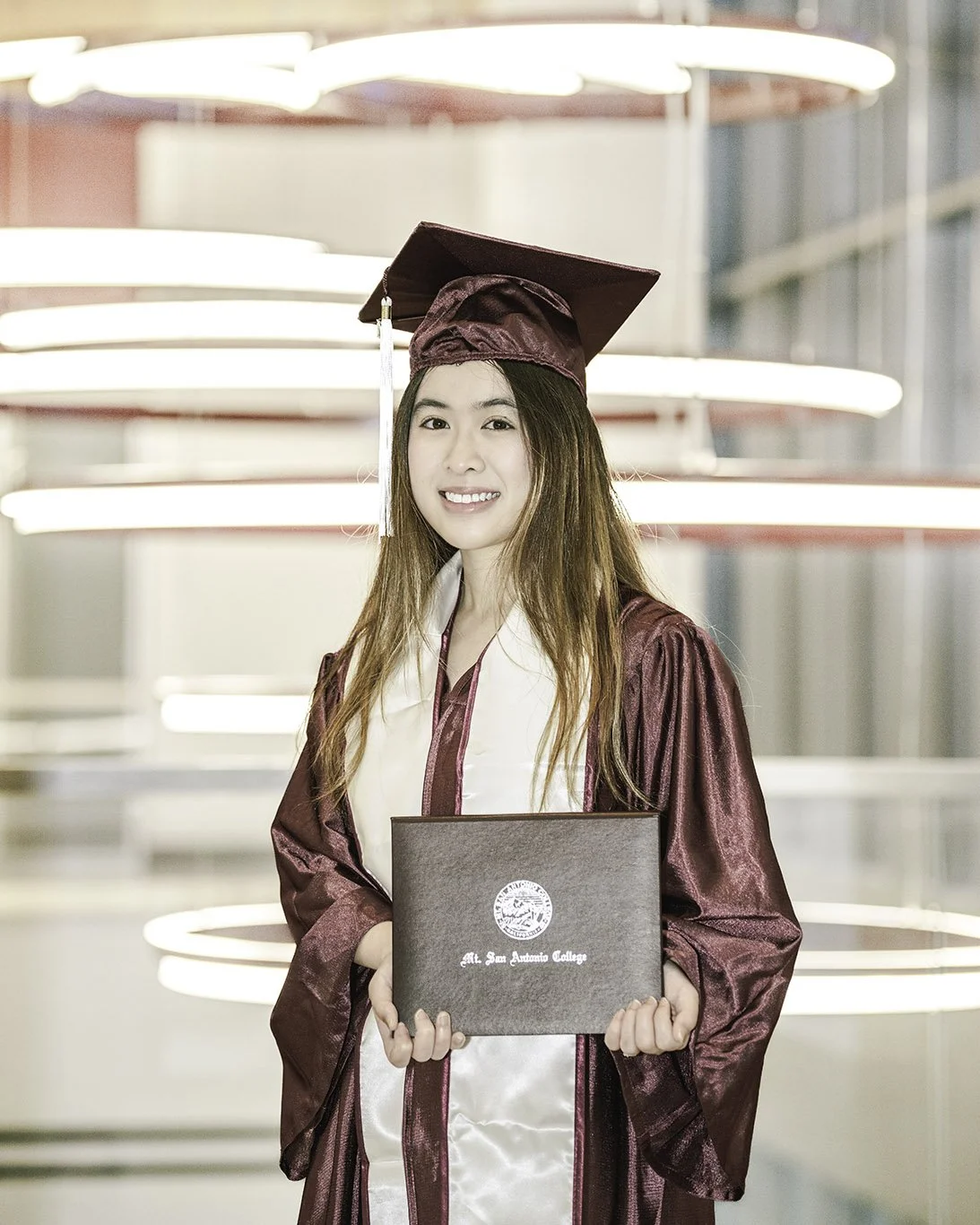 A young woman in a maroon graduation gown and cap holding a diploma in front of a modern, circular light fixture.