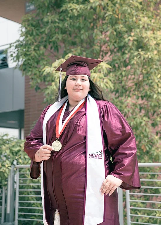 A young woman in a maroon graduation gown and cap, with a white sash, holding a gold medal, standing outdoors in front of green trees.