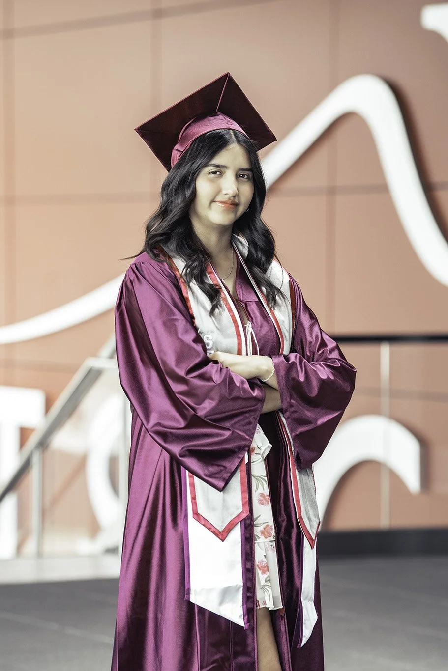 A young woman in a maroon graduation gown and cap stands with arms crossed, smiling at a graduation ceremony.