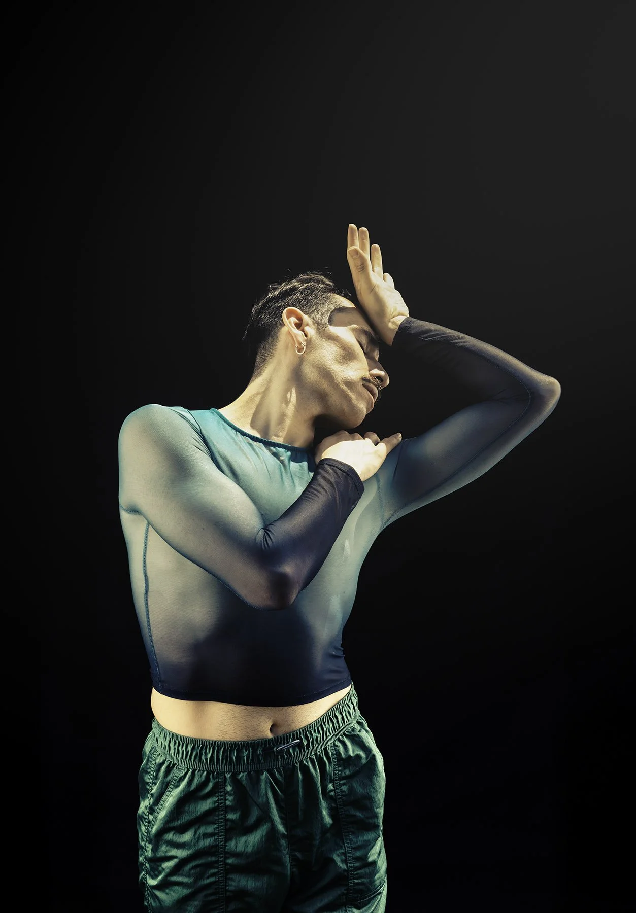 Young person with short dark hair performing a dance or expressive movement against a dark background, wearing sheer black and teal top and green shorts.