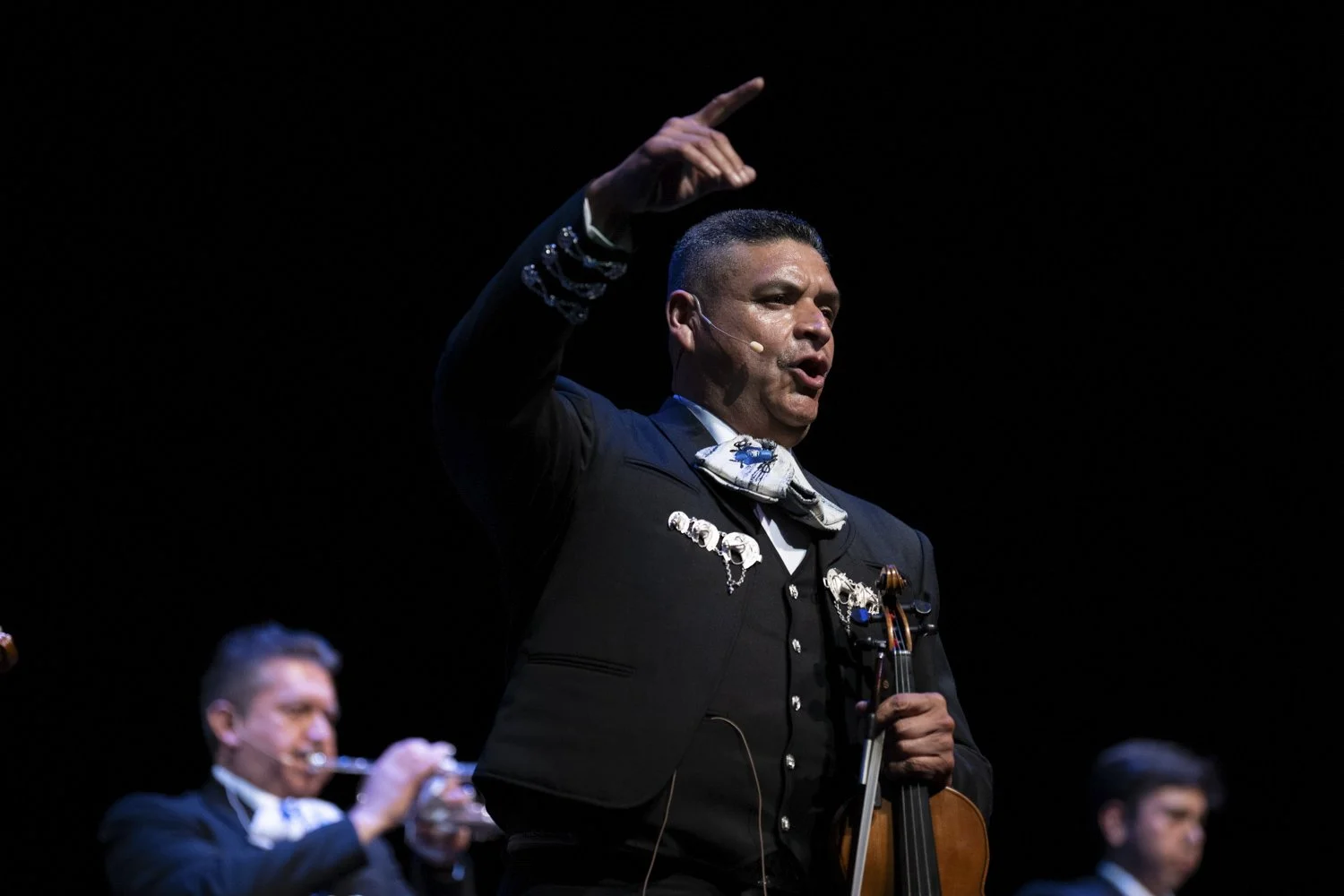 A man in a dark suit with a bolo tie wrapped around his neck holds a violin, raising his right hand with his index finger pointing upwards during a performance. Other musicians are blurred in the background.