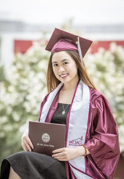 A young woman in a maroon graduation gown and cap, holding a diploma, smiling outdoors with a background of white flowers.